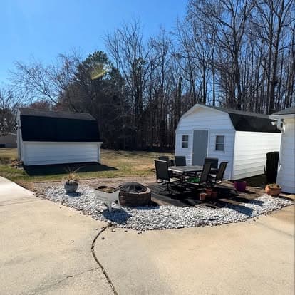 Two white sheds beside a stone patio with a fire pit and outdoor seating in a sunny backyard.