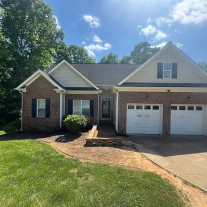 Brick house with dual garages, green lawn, and clear blue sky, surrounded by trees.